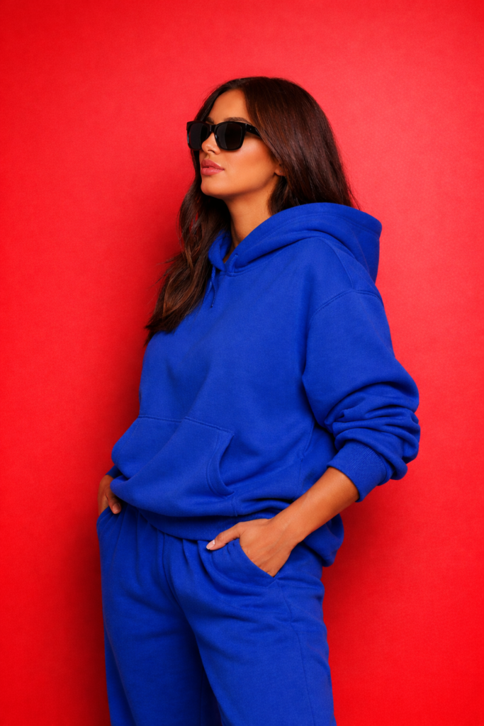 Chic woman styled in an elevated streetwear look featuring a purple hoodie and matching sweatpants, with long hair and sunglasses, posing confidently against a red background.