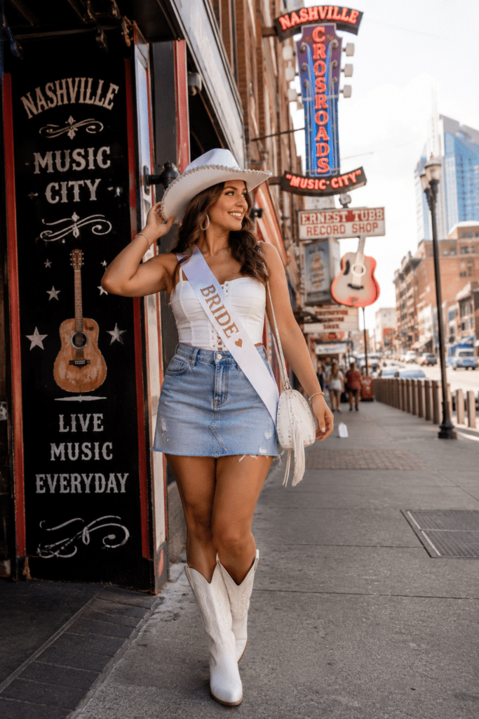 Woman wearing a white corset top, light-wash denim mini skirt, and white western boots walking on Broadway in Nashville with neon signs and a bridal sash.